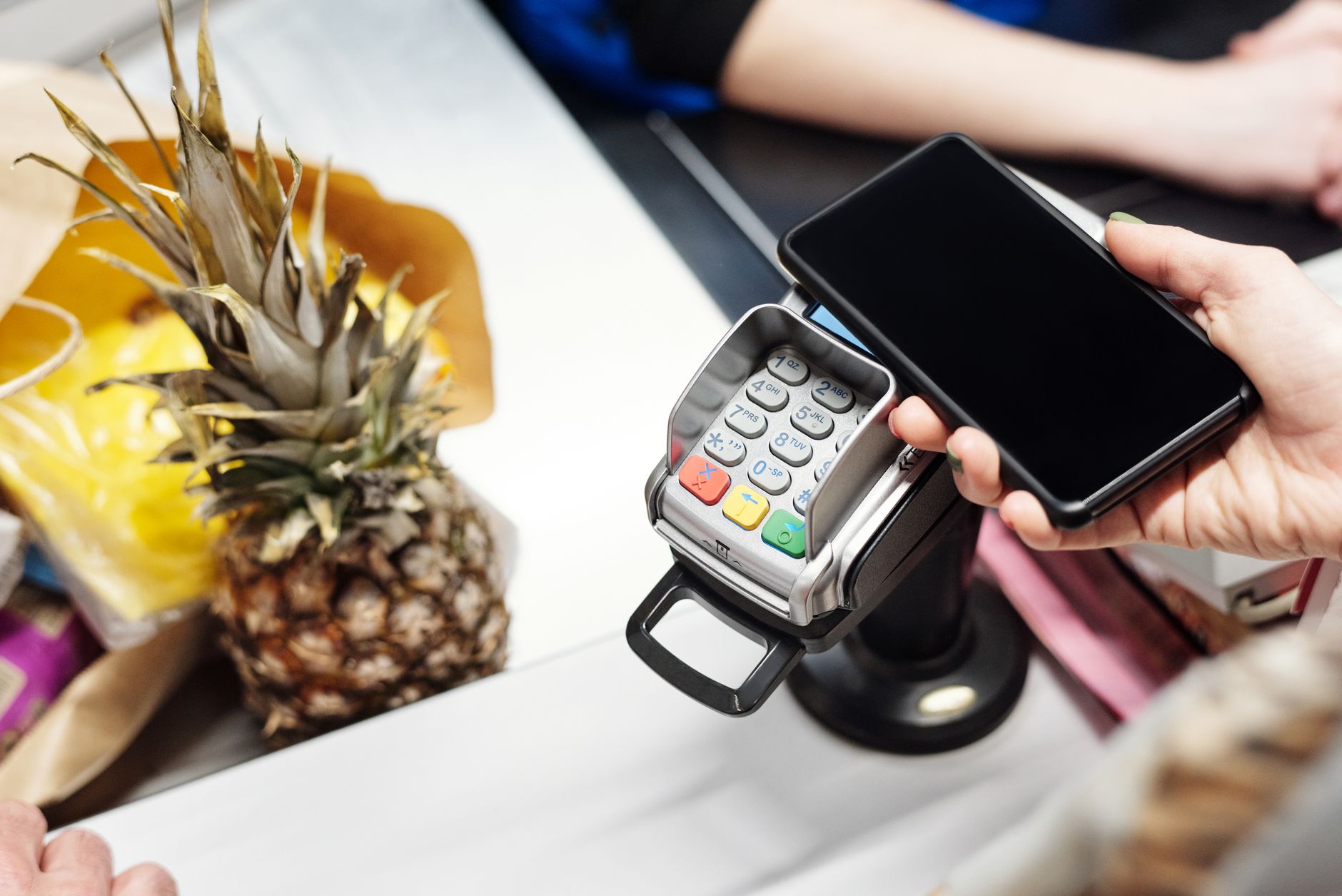 Gros plan d'un client utilisant un smartphone pour un paiement sans contact à la caisse d'un magasin avec un ananas sur le comptoir.
