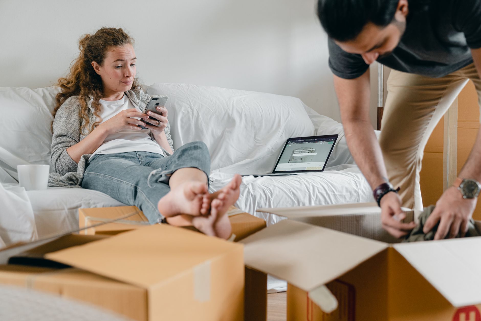 Femme pieds nus en tenue décontractée assise les jambes croisées sur un canapé avec un ordinateur portable ouvert tout en utilisant les réseaux sociaux sur son téléphone portable et son partenaire masculin récupérant des vêtements dans une boîte en carton.