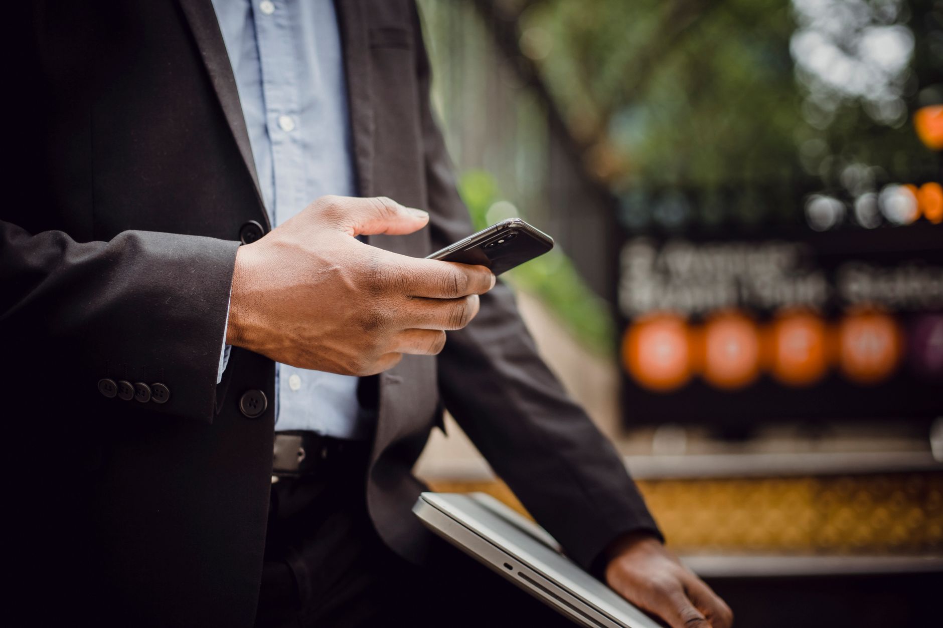 Homme afro-américain non reconnaissable en tenue formelle consultant un téléphone portable en se tenant avec un ordinateur portable dans la rue par une journée ensoleillée