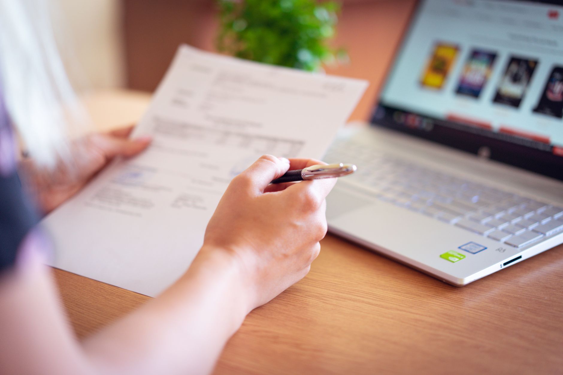 Une personne tient une feuille imprimée et un stylo devant un ordinateur portable ouvert sur un bureau en bois.